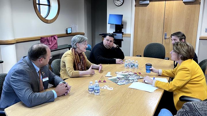 Five professionals are engaged in a discussion around a conference table, with documents and beverages in front of them. The setting includes a modern office environment.