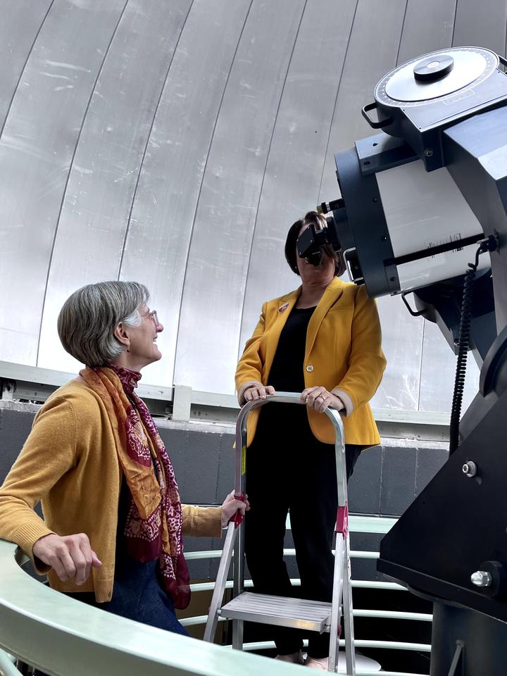 Two people at an observatory, one standing on a ladder looking through a large telescope, the other seated and looking up, both engaged in a conversation.