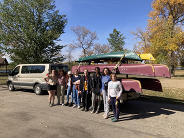 Eight people standing in front of a van and trailer loaded with canoes