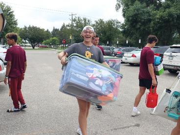 A white man wearing shorts, a grey t-shirt and a white baseball cap on backwards, carrying a large plastic tote during move-in day on campus. 