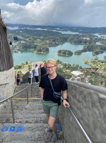 A blonde man with glasses, wearing a black t-shirt and green shorts, standing on a stairway overlooking the Medellin River in Medellin, Columbia