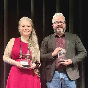 Two individuals holding awards on a stage, with one wearing a red dress and the other in a gray blazer, both smiling.