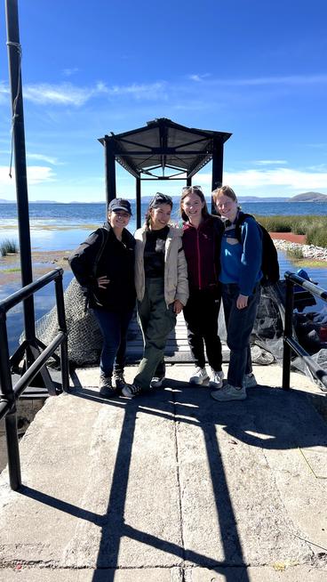 Four people standing together under a gazebo-like structure on a sunny lakeside pier, with mountains in the background.