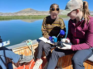 Two college researchers on a boat examining environmental samples with digital devices, with a scenic lake and hills in the background.