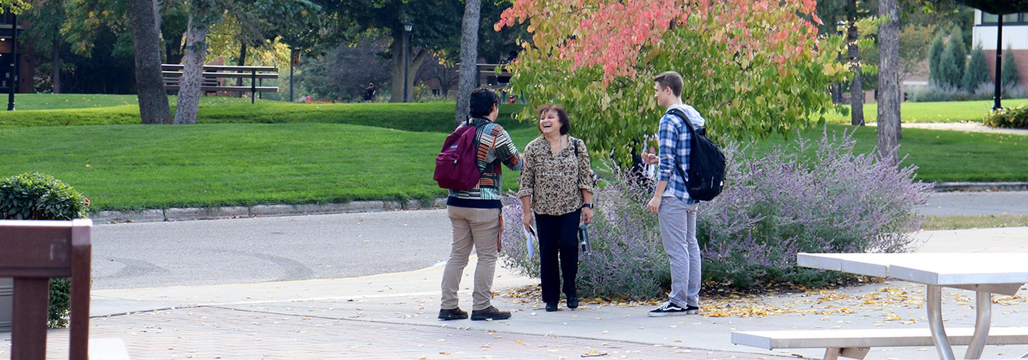 Two students talk with a faculty member on a sunny fall day on campus.