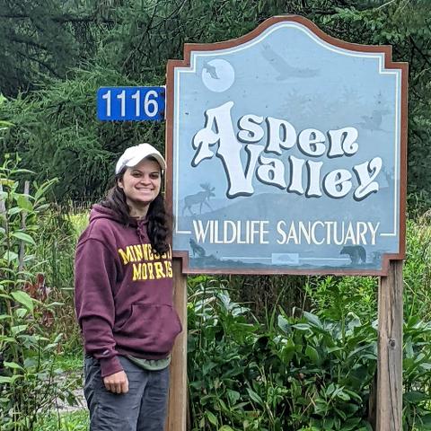 A man wearing a maroon Minnesota Morris sweatshirt and white baseball cap standing by a light blue sign that reads Aspen Valley Wildlife Sanctuary