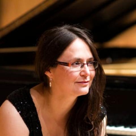 A woman with long dark hair and glasses wearing a short sleeved black dress, sitting at a black grand piano