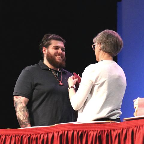 A young man with dark hair and beard wearing a black shirt receiving a medal from a woman with greying hair in a white sweater
