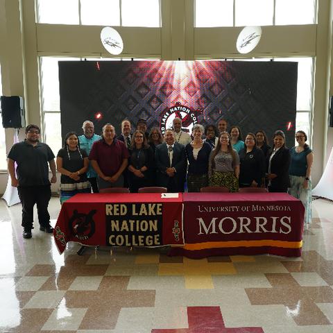 A group of 19 people standing behind a table with banners that read: "Red Lake Nation College" and "University of Minnesota Morris" 