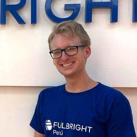 A smiling man with short blonde hair, wearing glasses and a blue t-shirt that reads Fulbright Peru