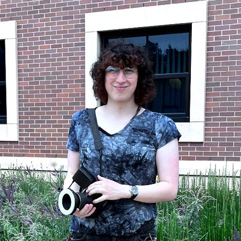 A person with dark curly hair, wearing a blue and black swirled t-shirt and holding a camera, standing by a flower garden in front of a brick building