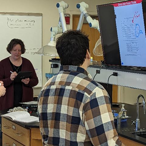 Two people in a chemistry lab.  A woman, the class professor, is holding an iPad.  The student has his back to the camera, and is looking at a TV monitor, which is displaying lab notes.  