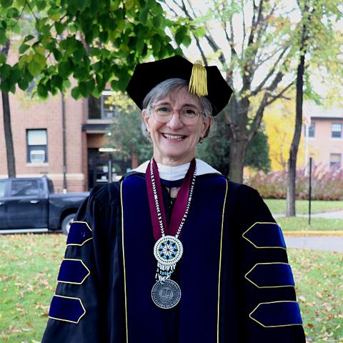 A white woman with gray hair and glasses wearing black academic regalia and two medallions