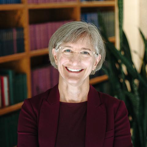 A smiling white woman with gray hair and glasses wearing a maroon shirt and blazer, standing in front of a bookcase. 