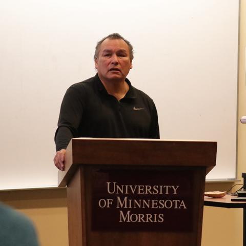 A Native American man with short grey hair, wearing a black long sleeved shirt, standing behind a podium with a sign that reads, University of Minnesota Morris. 