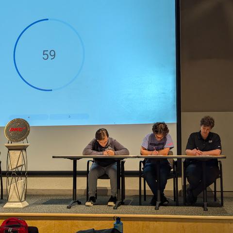 Three individuals seated at a desk in a lecture hall, focused on writing exams. A countdown timer is displayed on the screen behind them, showing 59 seconds.