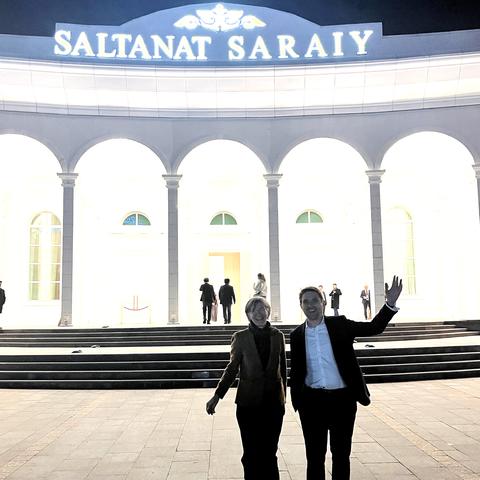 Two people smiling and standing in front of the Saltanat Sarayı at night, illuminated by a bright light. One person is waving.