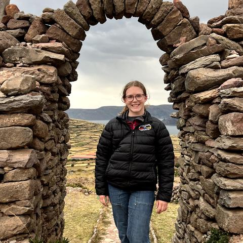 Maggie Landwer standing under an archway made of stones with a view of a terraced landscape in the background.