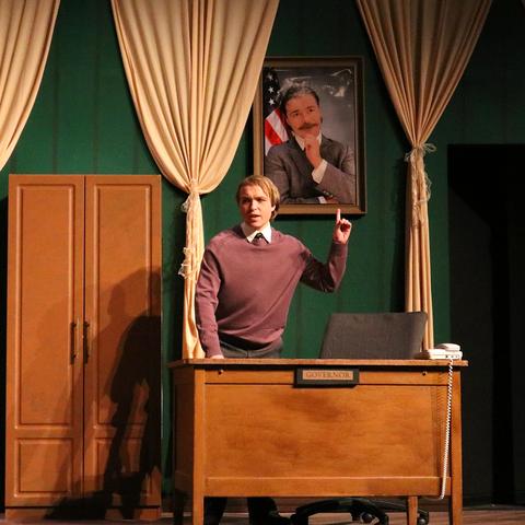 An actor on stage, standing by a desk, pointing at a portrait of Albert Einstein hanging on the wall in a room with green curtains and wooden doors.