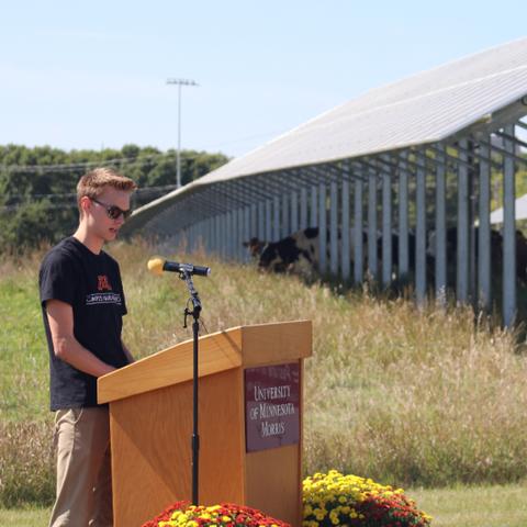 A college student standing at a podium at an outdoor event, with a field, cows and solar panels in the background. 