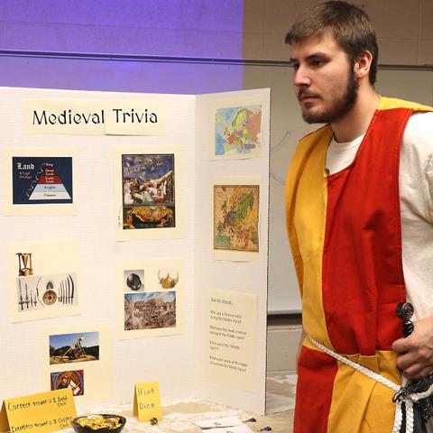 Person in medieval costume presenting a display board titled "Medieval Trivia" with various images and text about the medieval period.