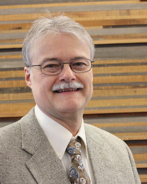 A photo of Steve Burks, a white man with grey hair and mustache, with glasses.  He is wearing a grey suit with a white shirt and multi-colored tie. 